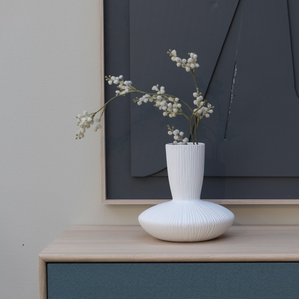 A white ribbed vase with delicate, white floral branches is placed on a light wooden surface. The background features a textured dark artwork framed against a light wall.