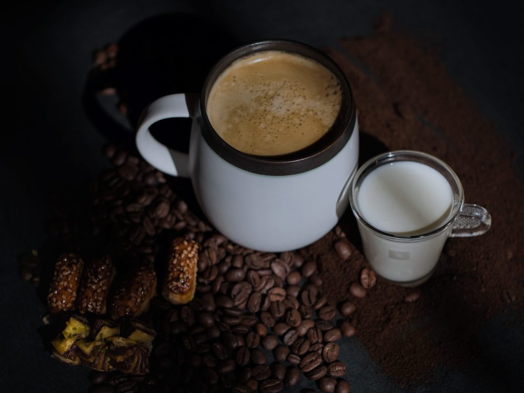 A white mug filled with coffee, accompanied by a small glass of milk and some sesame-studded sweets, arranged on a dark surface scattered with coffee beans and ground coffee.