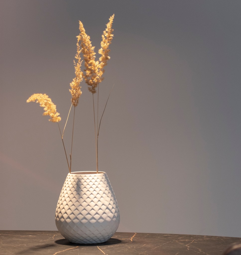 A modern, textured white vase with three tall, dried pampas grass stems, set against a soft gray background and a dark surface.