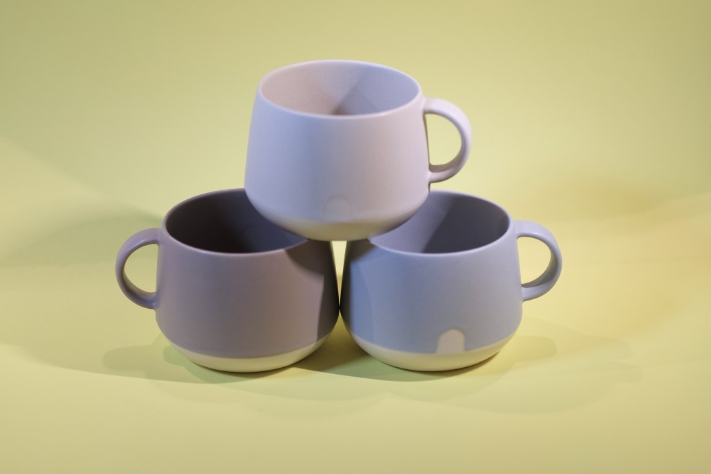 Three ceramic cups in different shades of purple and blue, stacked together on a light yellow background.